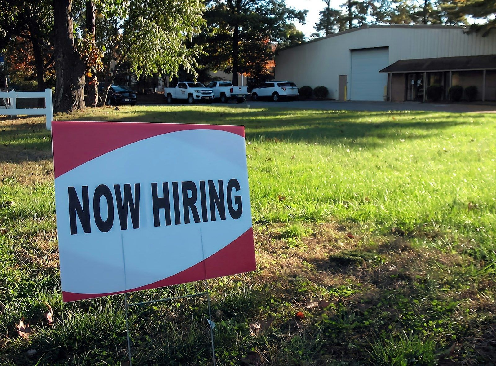 A Now Hiring sign on the lawn in front of a shop, with work trucks parked outside.