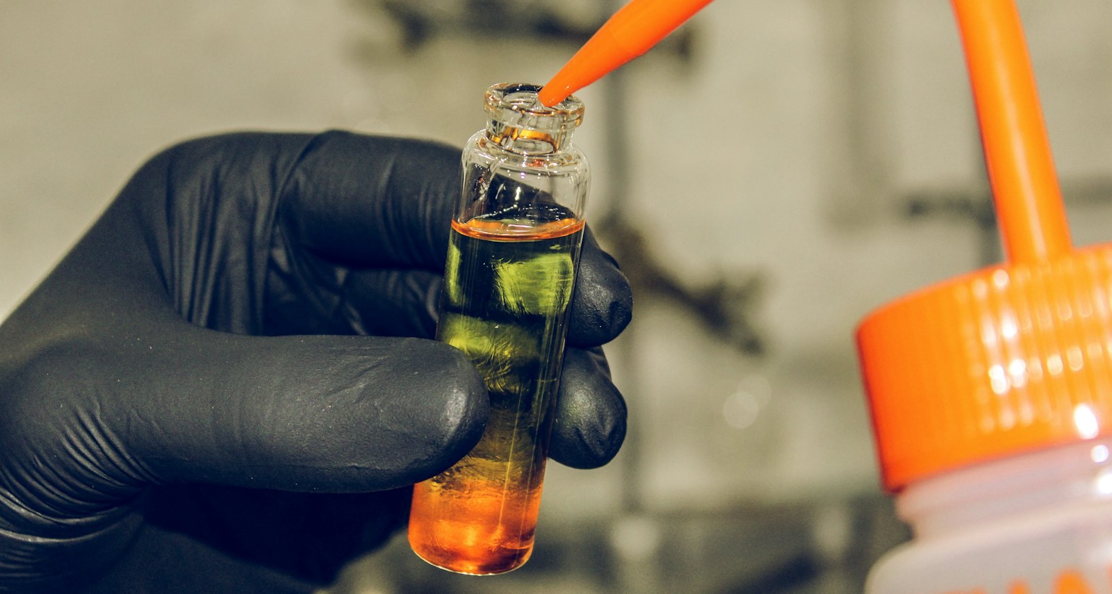 A gloved hand holding a sample vial while a dropper adds reagent.