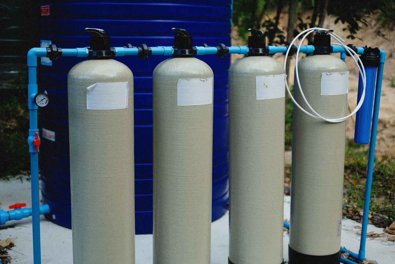 Four filter tanks plumbed in a row with blue pipe and a pressure gauge.