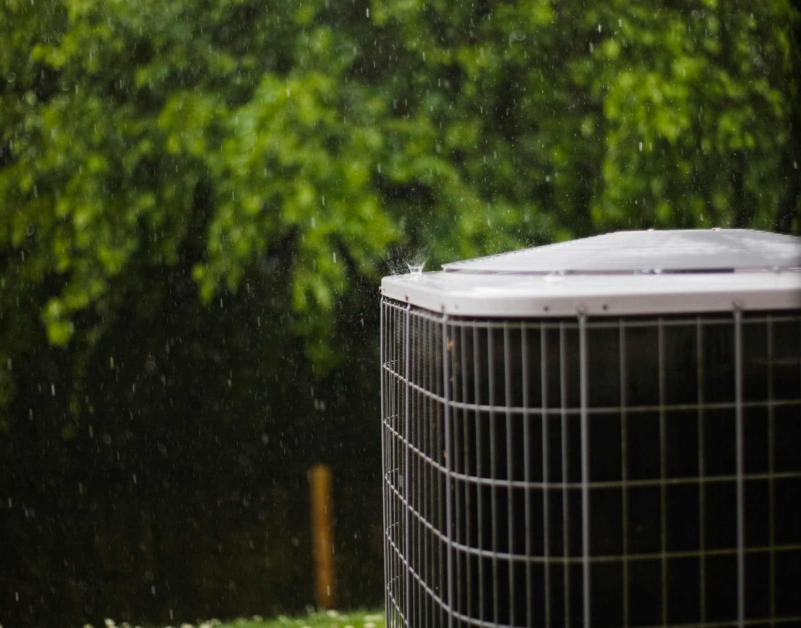 An outdoor condenser unit standing in the rain, with trees behind it.