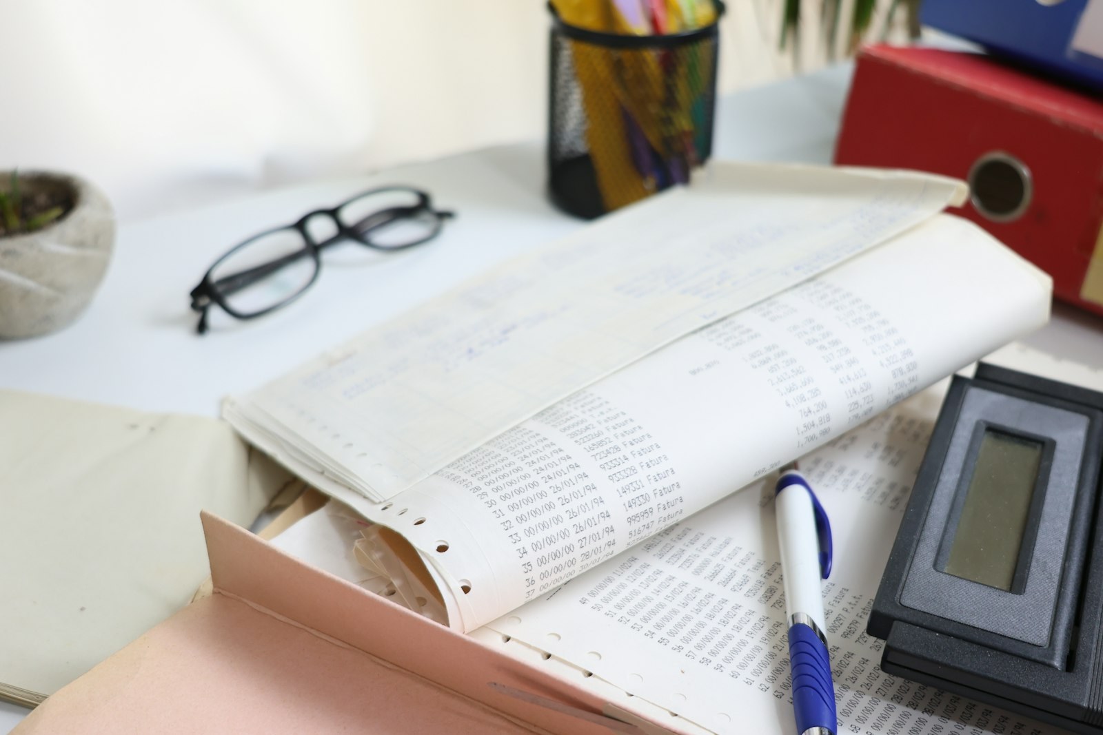 Printed ledgers, a pen, a calculator and a pair of glasses on a desk.