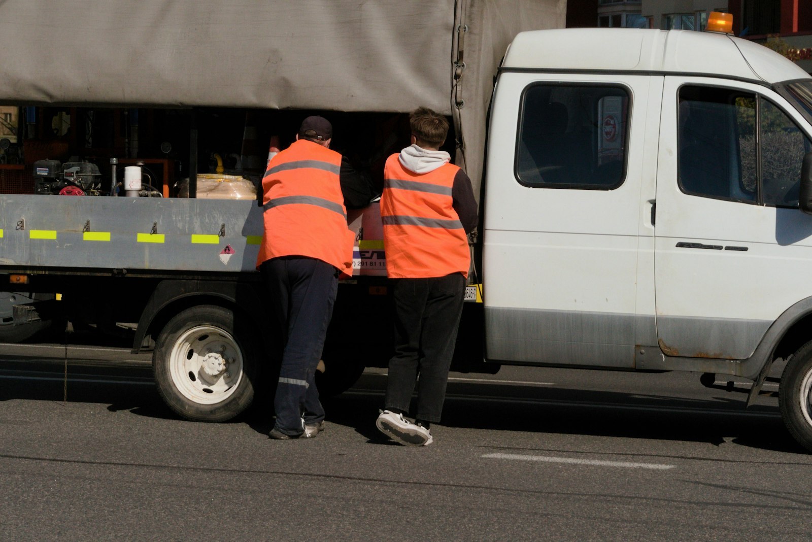 Two workers in orange vests reaching into the side of a white truck.