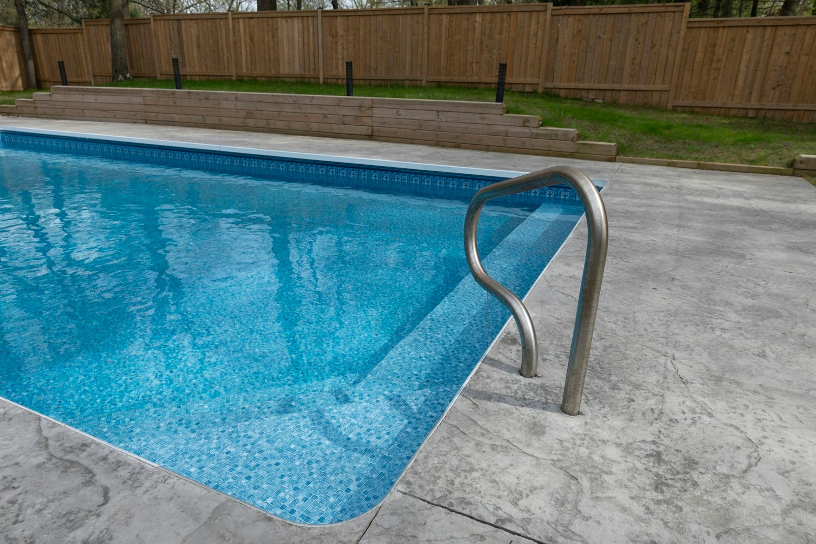 The corner of a backyard pool with a steel handrail and a wooden fence behind it.