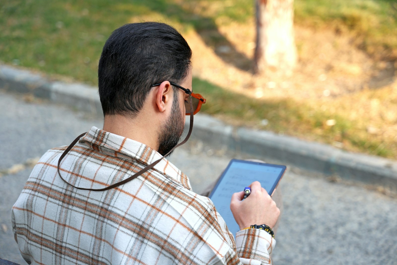 A man in a plaid shirt working on a tablet outdoors, seen from behind.