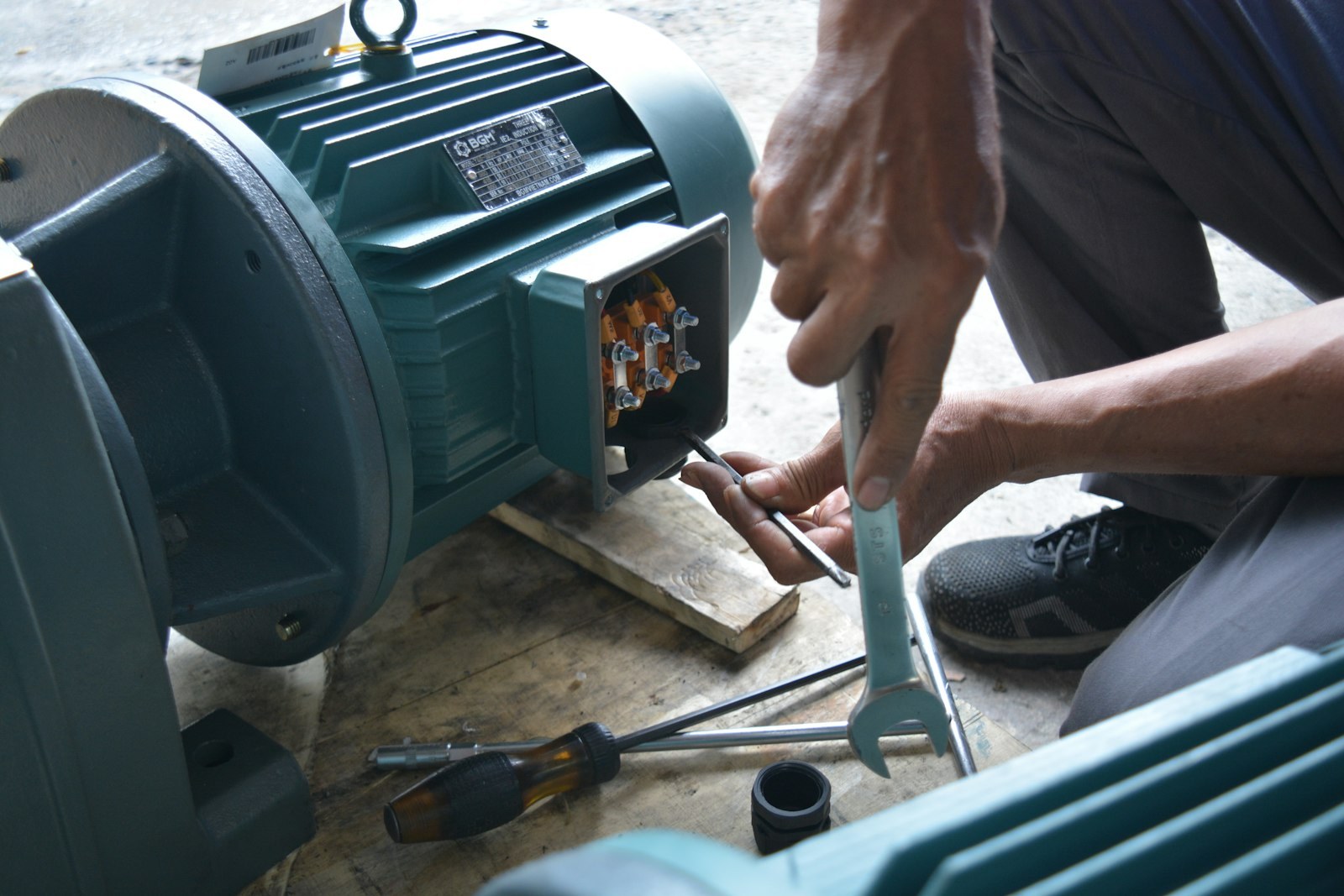 A technician working on the wiring box of an electric motor, with a wrench and screwdrivers on the ground.