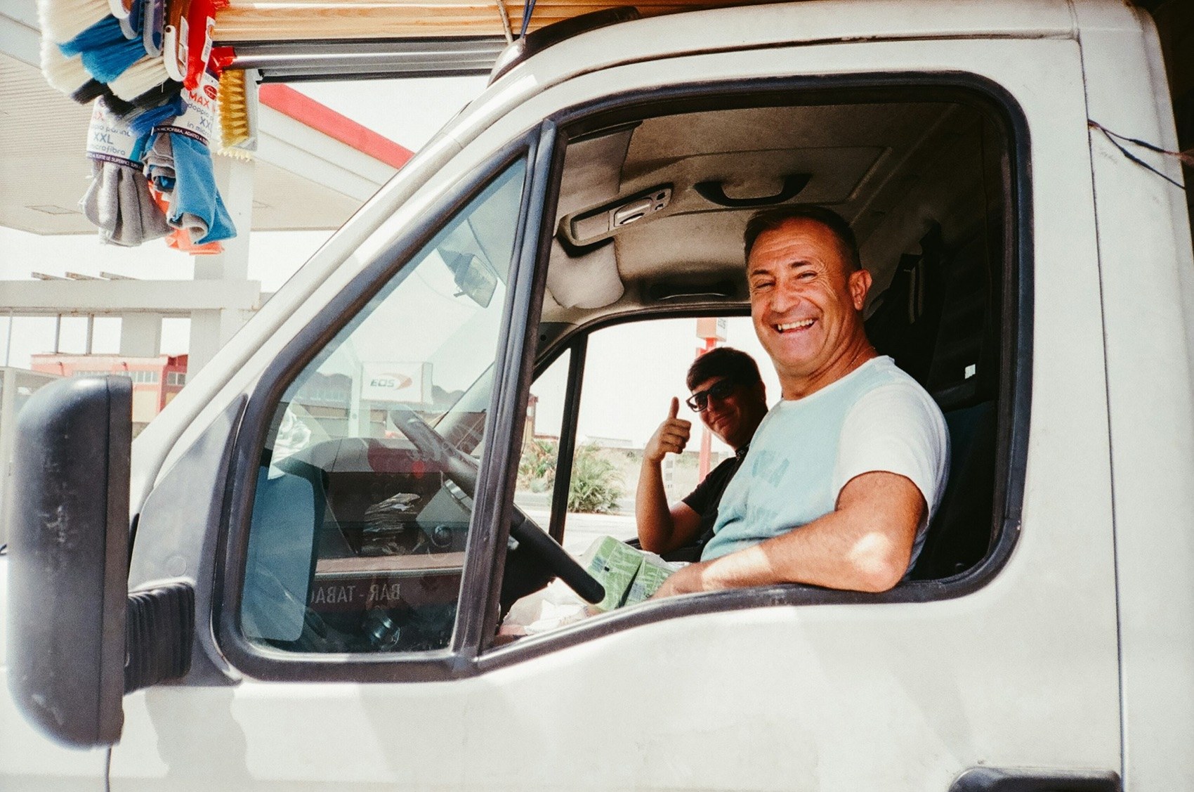 Two men smiling from the cab of a white work van, one giving a thumbs up.