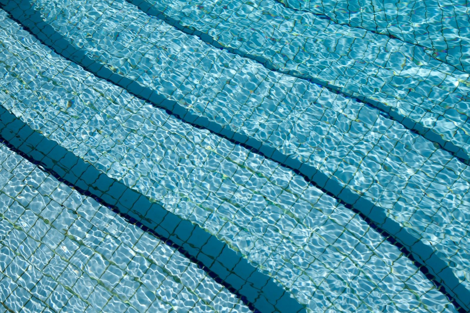 The tiled floor of a swimming pool with dark lane lines, seen through rippling water.