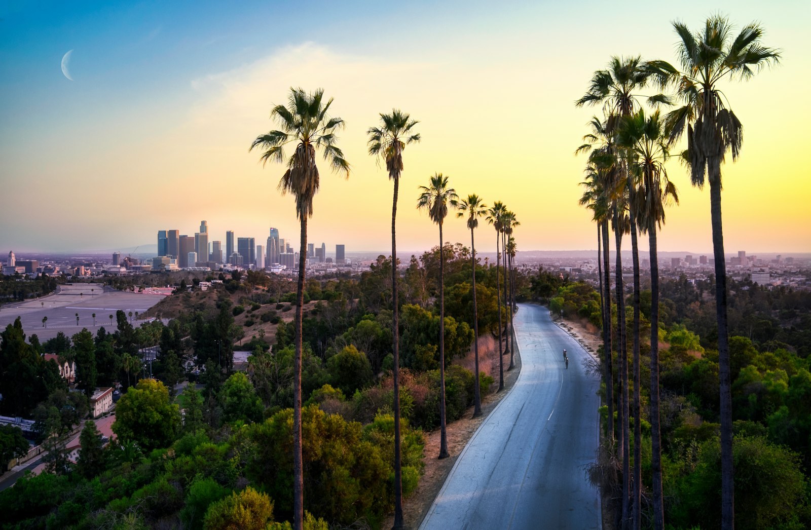 A palm-lined road in Los Angeles with the downtown skyline in the distance at sunset.